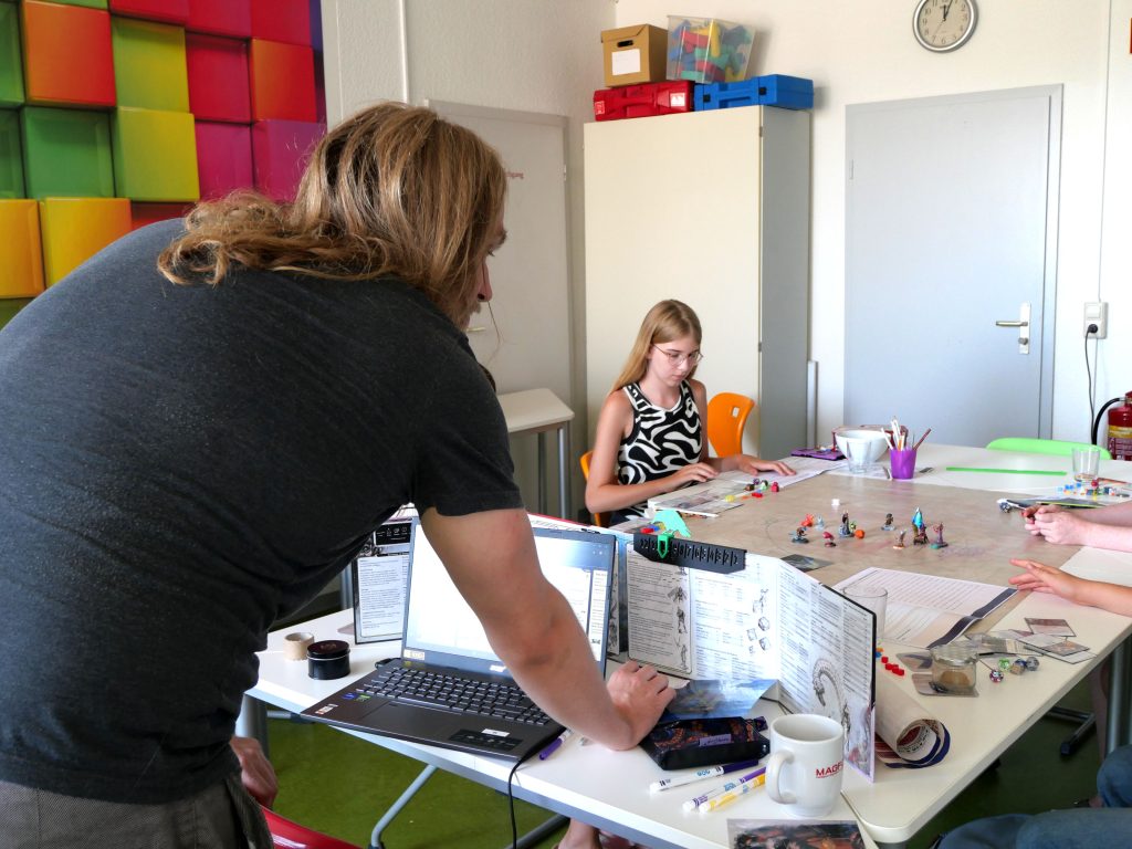 Ein Mann steht mit dem RÜcken zur Kamera, im Hintergund sitzt ein Mädchen an einem Tisch. Auf dem Tisch liegen Spielfiguren und Würfel.
