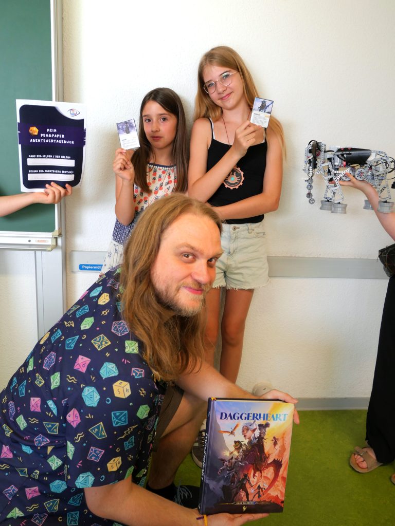 Drei Menschen mit Spielkarten und einem Buch mit dem Titel "Daggerheart", schauen in die Kamera.